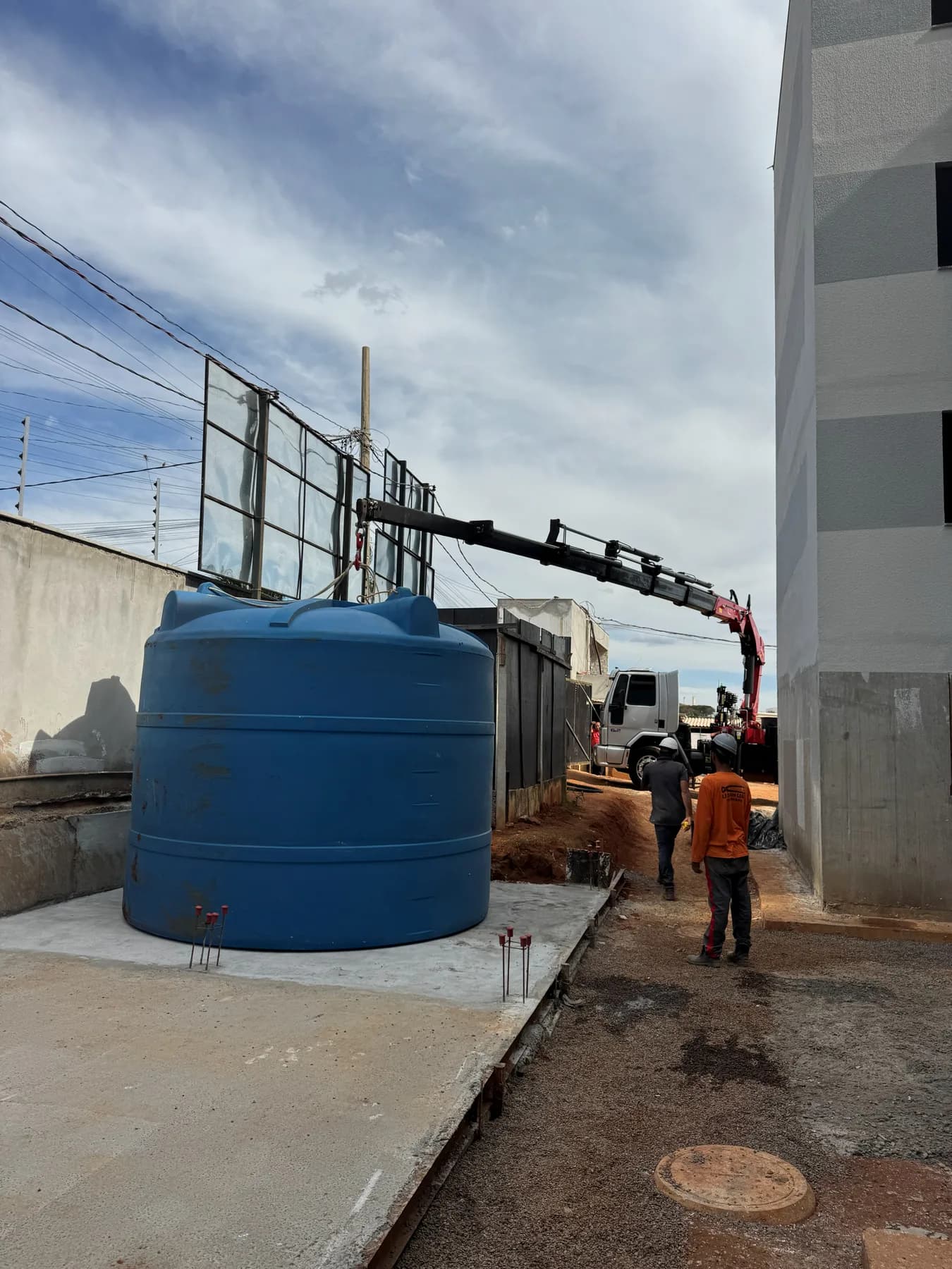 Caminhão munck com a lança estendida posicionando uma caixa d'água azul de grande porte sobre a base de concreto, ao lado de um prédio em obra